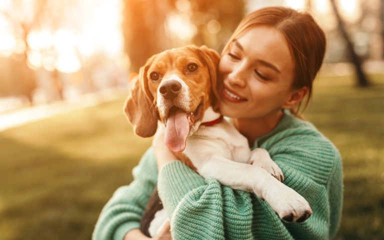 Een jonge vrouw houdt een blije hond in haar armen terwijl ze samen buiten zitten, omringd door warm, goudkleurig zonlicht.